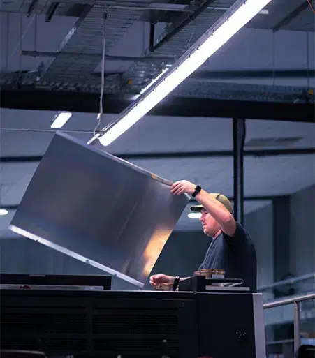 Service-home-2 Man in a cap lifts a large metal sheet inside an industrial printing or manufacturing facility with overhead lighting.