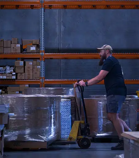 Service-home-4 Bearded man wearing a cap pushing a pallet jack with wrapped pallets in a warehouse with stacked boxes on shelves.