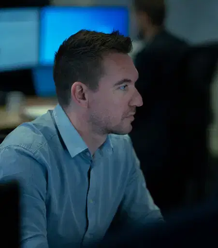 Service-home-5 Man in a blue button-up shirt focused on work, sitting in front of computer screens in a dimly lit office environment.