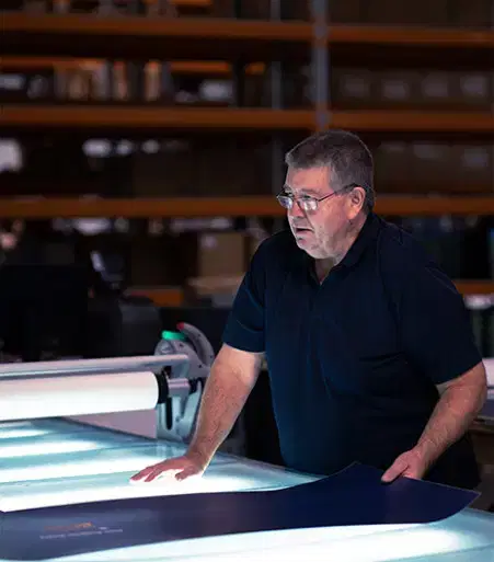Service-home-7 Man wearing glasses inspecting a large printed sheet on a lit table in a warehouse or print production environment.
