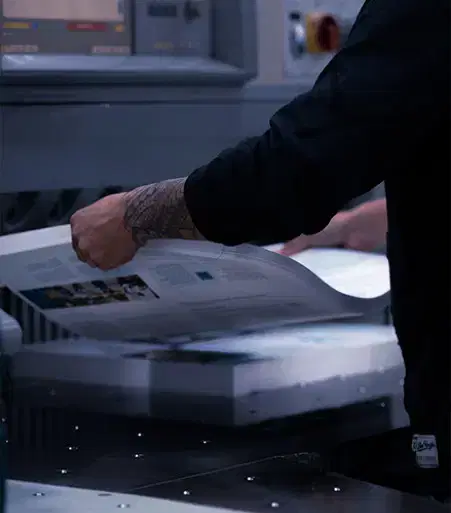 Service-home-9 A person with tattooed arms handles freshly printed sheets being ejected from a commercial printing machine.