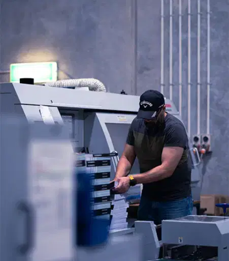Service-managed-Service Man in a black cap operating a large industrial printing machine inside a dimly lit modern warehouse or print shop.