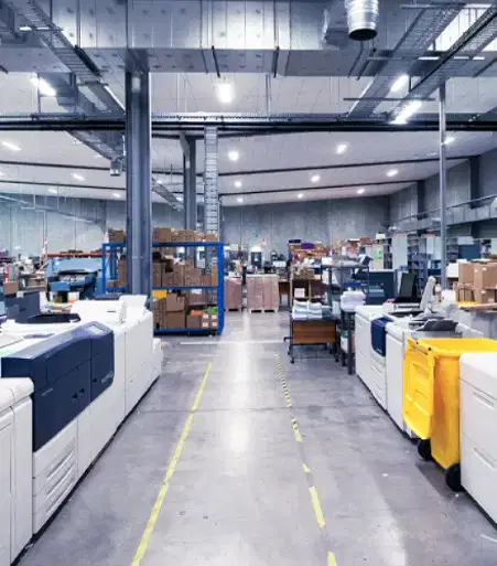 Services-Inovation A large industrial printing warehouse with rows of printers, stacks of paper, and organized shelves under bright lights.