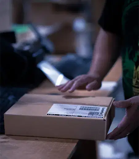 Services-Inventory Person’s hands holding a cardboard box with a shipping label on a table in a dimly lit workspace or warehouse environment.