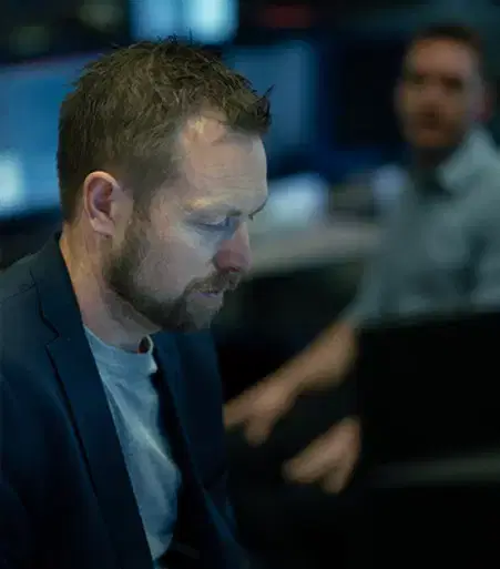 Services-Online Focused man with beard and blazer working intently at a computer desk in a dimly lit office with blurred colleague behind.