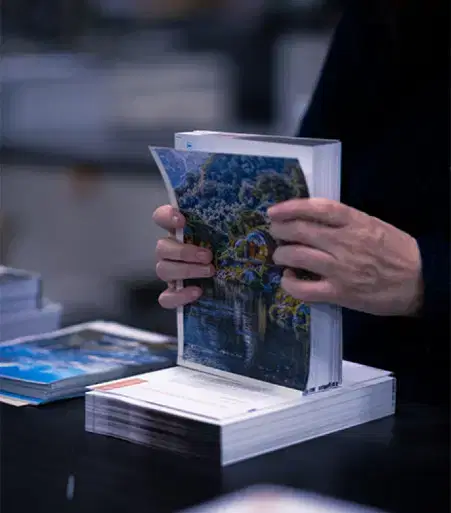 Services-Printing-1 Hands flipping through a thick booklet with colorful images, stacks of similar booklets on a dark table in a print shop.