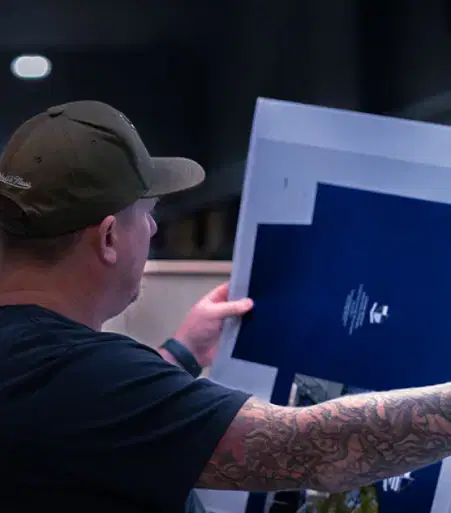 Services-Web Tattooed man wearing a cap inspects printed sheets in a dimly lit commercial printing facility with focused attention.