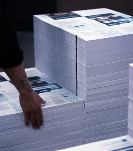 Services-complex-order A person’s hand with tattoo organizes stacks of freshly printed paper sheets in a professional printing facility.
